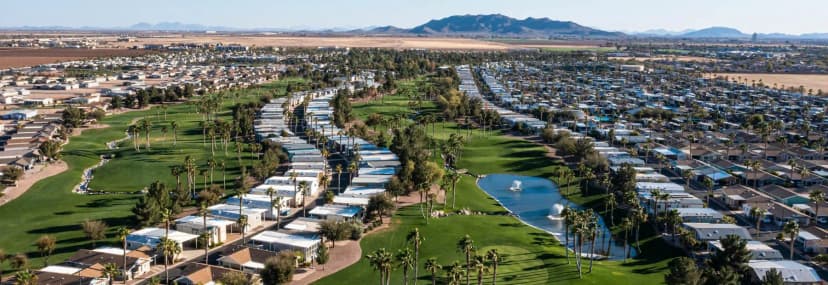 Aerial view of a desert residential community with rows of homes bordering a golf course with a fountain pond and palm trees, with mountains in the distance.